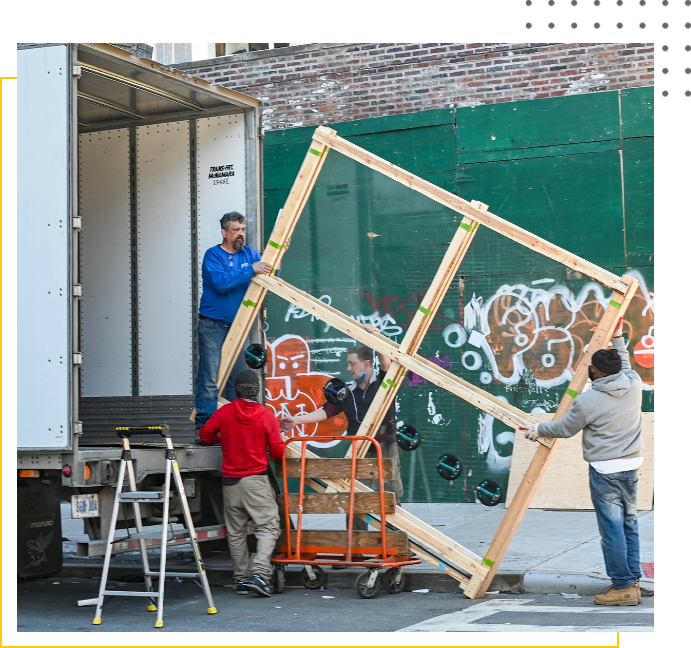 Three workers load a large, framed object into a truck parked by a graffiti-covered wall.